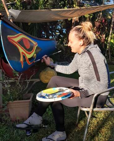 Eine Frau sitzt draußen auf einem Metallstuhl und bemalt ein blaues Kanu mit einem bunten ägyptischen Horus-Auge-Motiv, umgeben von üppigem Grün und Sonnenschein.