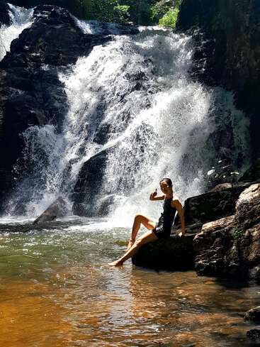 Eine Frau sitzt auf einem Felsen neben einem rauschenden Wasserfall inmitten einer üppigen Naturlandschaft. Sonnenlicht taucht die Szene in ein warmes Licht und unterstreicht die erfrischende, ruhige Atmosphäre.