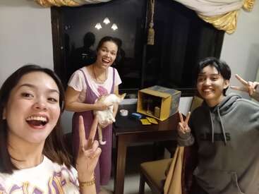 Tres personas sonrientes posan en un interior, dos de ellas mostrando signos de la paz. Una sostiene un gato blanco. Detrás, una mesa con una caja amarilla y cartas. Ambiente nocturno.