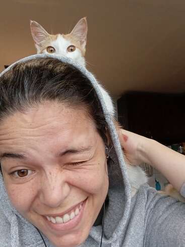 Eine Person in einem grauen Kapuzenpulli zwinkert und lächelt in die Kamera, während eine neugierige orange-weiße Katze von hinten über ihre Kapuze lugt.