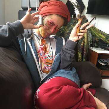 Una persona sonriente con la cara pintada de colores está sentada en un sofá, lleva un pañuelo en la cabeza, sudadera con capucha y pantalones rojos, y sostiene un pequeño recipiente y un pincel.