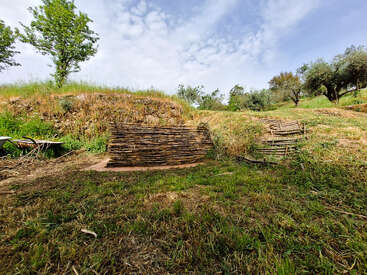 A rustic landscape with grassy hills, scattered trees, and two woven wooden structures. The sky is partly cloudy, creating a peaceful and natural rural atmosphere.