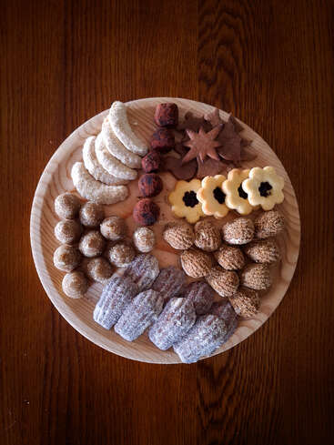 A wooden plate holds a beautifully arranged assortment of cookies and pastries, including crescents, truffles, stars, jam-filled, nut-shaped, and powdered treats on a wood table.
