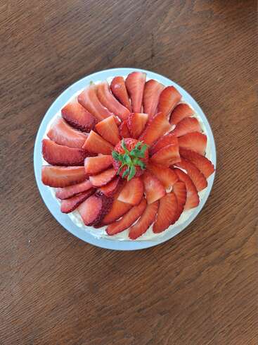 This image shows a beautifully decorated cake topped with neatly arranged, fresh strawberry slices in a circular pattern, sitting on a white plate on a wooden table.