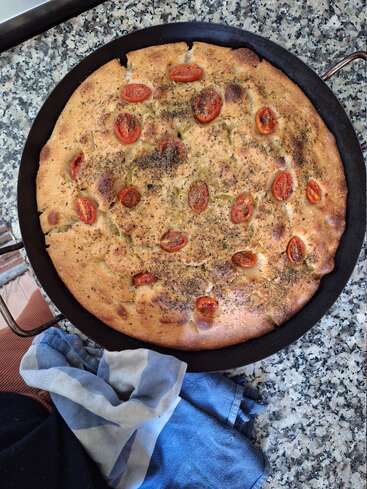 A freshly baked round focaccia bread topped with halved cherry tomatoes and herbs, sits in a black pan on a speckled countertop, with a kitchen towel nearby.