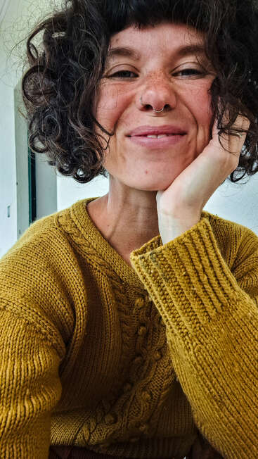 Eine Frau mit lockigem dunklem Haar, einem Nasenring und Sommersprossen lächelt herzlich. Sie trägt einen senfgelben Strickpullover und stützt ihr Kinn auf die Hand.