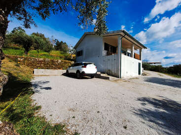 Um carro branco está estacionado ao lado de uma casa de dois andares com uma varanda coberta. A região é rural, com vegetação, árvores, céu azul e nuvens.