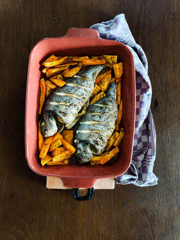 Uma travessa de cerâmica contém dois peixes assados inteiros, rodeados por fatias de batata-doce temperadas. A travessa está apoiada sobre uma superfície de madeira, com um pano xadrez ao lado.