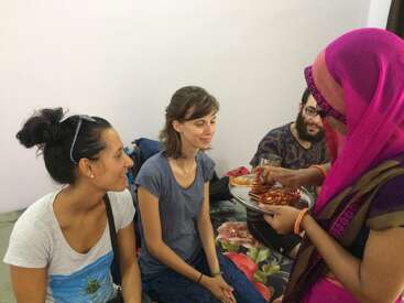 La imagen muestra a un grupo de cuatro individuos reunidos en torno a una mesa, con dos mujeres y un hombre sentados en una cama, mientras una mujer con un pañuelo rosa en la cabeza sirve comida de un plato.