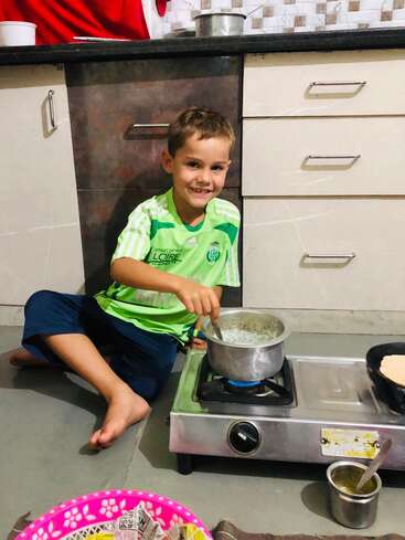 Un niño está sentado en el suelo de una cocina, removiendo una olla en el fogón, con una camisa verde y unos pantalones azules, y una sonrisa en la cara.