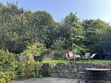 Um jardim com árvores e arbustos verdejantes, paredes de pedra, uma área de pátio, cadeiras de madeira ao ar livre, uma mesa e um trampolim sob um céu claro.