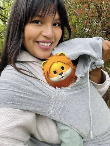 Das Bild zeigt eine Frau mit langen dunklen Haaren, die lächelt und einen hellgrauen Kapuzenpullover trägt, aus dessen Kapuze ein Cartoon-Löwe herausschaut.