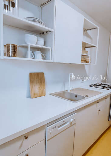 Das Bild zeigt eine moderne Küche mit weißen Schränken, Arbeitsplatten und Regalen, mit Spüle, Geschirrspüler, Herd und Schneidebrett vor einer sauberen weißen Wand.