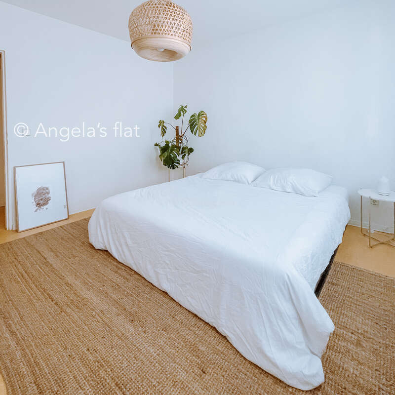 Das Bild zeigt ein ruhiges Schlafzimmer mit einem weißen Bett, einem beigen Teppich und einer Pflanze, mit einer Korbleuchte und einem gerahmten Bild an der Wand.