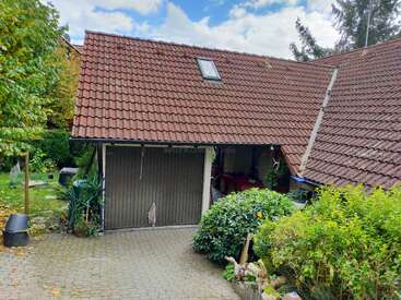 A house with a sloped red-tiled roof, small skylight, attached garage, lush greenery, bushes, and trees, set under a partly cloudy blue sky.