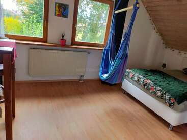Bright room with large windows, wooden floor, bed with floral blanket, blue hanging hammock chair, small table, artwork, and a potted plant by the radiator.