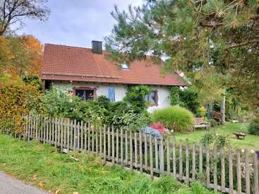 A cozy house with a red tiled roof sits behind a wooden picket fence, surrounded by lush green plants, trees, and a well-tended garden.