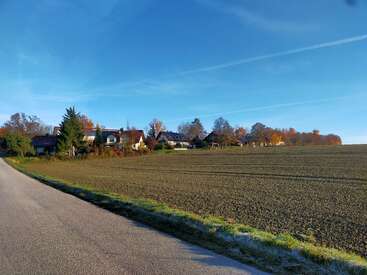Eine ruhige Landstraße führt neben einem frisch gepflügten Feld zu Häusern, die von herbstlichen Bäumen unter einem klaren, blauen Himmel mit schwachen Kondensstreifen umgeben sind.