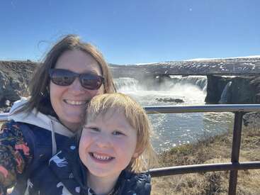 Eine Frau und ein Kind lächeln für ein Selfie vor einem malerischen Wasserfall unter strahlend blauem Himmel und genießen gemeinsam einen sonnigen, fröhlichen Tag im Freien.