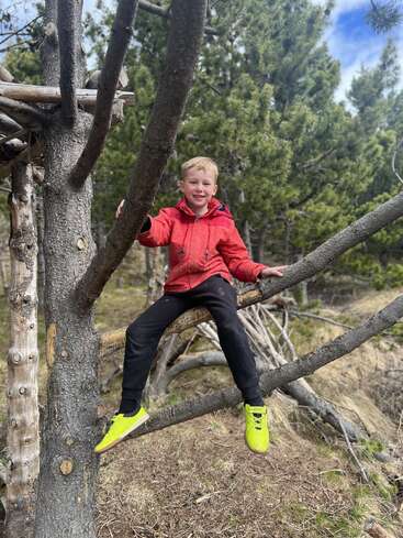 Ein lächelnder Junge in einer roten Jacke und leuchtend gelben Schuhen sitzt auf einem Ast im Wald, umgeben von hohen Kiefern und der Natur.