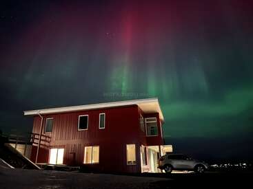 Ein gemütliches rotes Haus mit beleuchteten Fenstern steht unter einem Nachthimmel, der von leuchtend grünen und rosa Nordlichtern erhellt wird, daneben steht ein Auto. Eine zauberhafte Atmosphäre.