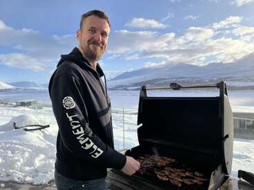 Ein Mann in einem schwarzen Kapuzenpulli grillt draußen Fleisch, umgeben von schneebedeckten Bergen unter einem strahlenden, teilweise bewölkten Himmel. Er lächelt und genießt das Wintergrillen.