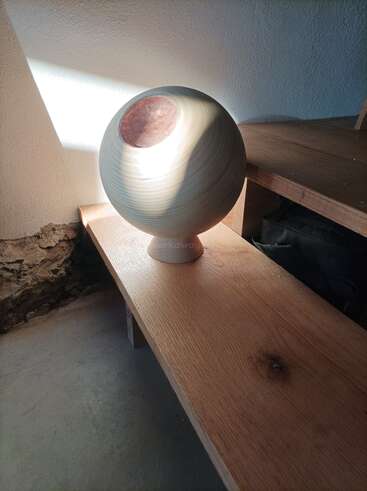 Sobre un banco de madera descansa un jarrón redondo de cerámica de color claro y boca ancha. La luz del sol lo ilumina, proyectando sombras sobre la pared y la superficie de madera.
