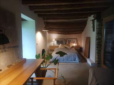 Este acogedor dormitorio cuenta con vigas de madera de estilo rústico, una cama bien hecha con cojines, un escritorio de madera, plantas de interior, una iluminación tenue, detalles en piedra y una decoración minimalista.