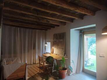 Esta imagen muestra una habitación acogedora y rústica con vigas de madera a la vista en el techo, un escritorio con una silla, una planta en maceta, una cama, un gran ventanal, cortinas y una iluminación tenue.