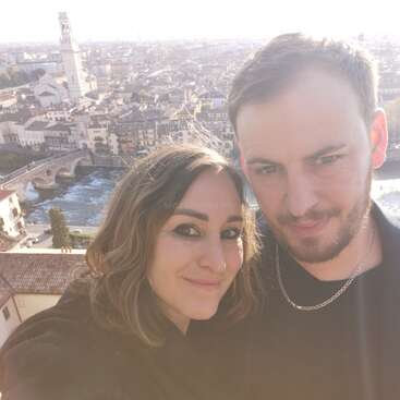 The image shows a selfie of a man and woman with a cityscape in the background, featuring a river and bridge, and a church steeple.