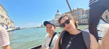 Une femme et un garçon sourient en montant sur une gondole à Venise, en Italie, avec des bâtiments historiques et un dôme visibles en arrière-plan. Journée ensoleillée.