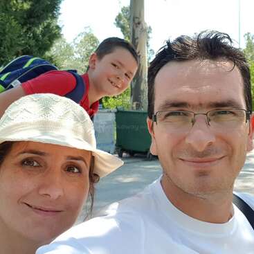 Une famille souriante prend un selfie joyeux en plein air. L'homme porte des lunettes, la femme un chapeau blanc et un jeune garçon en rouge pose derrière eux.