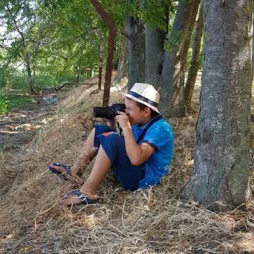 Un jeune garçon portant un chapeau et des vêtements bleus est assis sur de l'herbe sèche près d'arbres, tenant un appareil photo et photographiant la nature, profitant d'une paisible journée d'été en plein air.