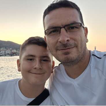 Un père et son fils posent pour un selfie au bord de la mer au coucher du soleil, tous deux souriants, avec des maisons, un voilier et de douces collines visibles en arrière-plan.