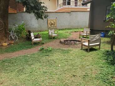 A cozy outdoor seating area with four wooden chairs around a stone fire pit, surrounded by grass, trees, a bicycle, and a painted wall sign.