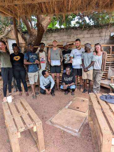 A group of twelve people pose together under a thatched shelter. Two individuals hold certificates, while others smile and crouch, suggesting a celebratory or graduation event.