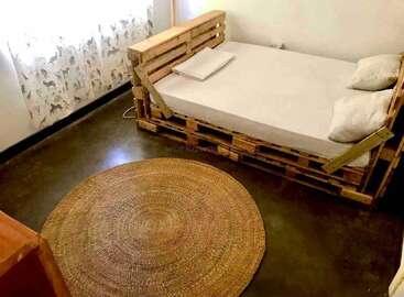 A minimalist bedroom features a bed made from wooden pallets, white bedding, animal-print curtains, a round woven jute rug, and polished dark concrete floors. Cozy, simple decor.