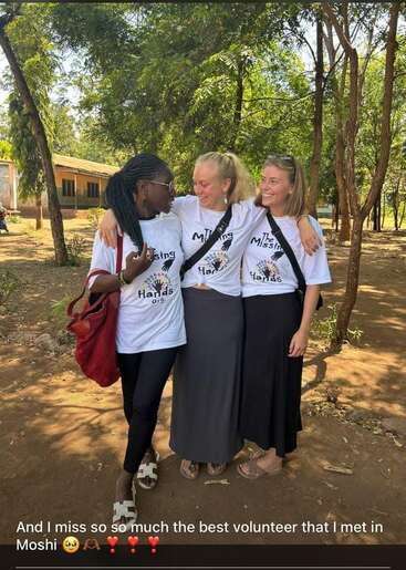Três mulheres estão do lado de fora, de braços dados, sorrindo calorosamente. Todas vestem camisetas da “The Missing Hands Org” e estão rodeadas por árvores. Amizade genuína e felicidade irradiam neste momento memorável de voluntariado.