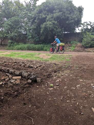 A person rides a loaded touring bicycle along a dirt path, surrounded by greenery and trees, with stone piles and dry soil in the foreground.