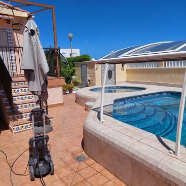 A backyard featuring a clean swimming pool and hot tub, tiled patio, decorative stairs, umbrella, cleaning equipment, and clear blue skies on a sunny day.