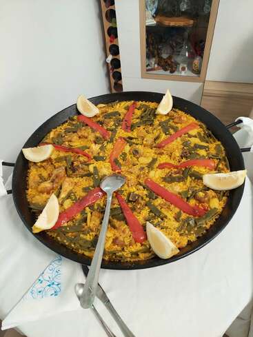 A large traditional paella in a black pan, garnished with lemon wedges and red pepper strips, sits on a white tablecloth, ready to be served.