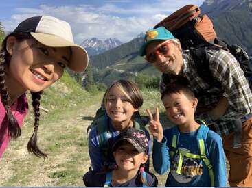 La imagen muestra a una familia de cinco miembros posando para una foto en un entorno montañoso. La mujer de la izquierda lleva una gorra de béisbol y el hombre de la derecha una gran mochila.