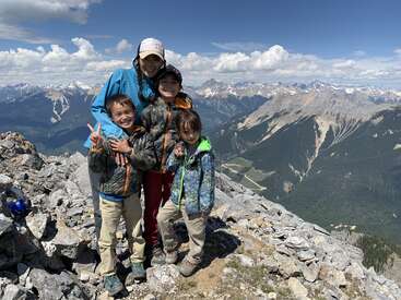 La imagen muestra a una mujer y tres niños posando en la cima de una montaña rocosa, con una impresionante vista de montañas y valles de fondo bajo un cielo azul con nubes.