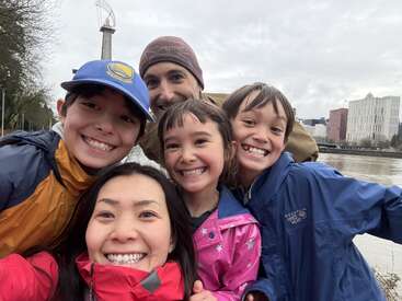 A happy family of five, bundled in colorful jackets, smiles brightly for a selfie outdoors on a cloudy day, with an abstract structure in the background.
