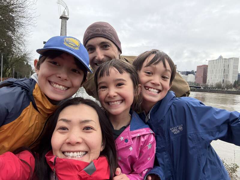 Una feliz familia de cinco miembros, abrigados con chaquetas de colores vivos, sonríe alegremente para hacerse un selfi al aire libre en un día nublado, con una estructura abstracta al fondo.