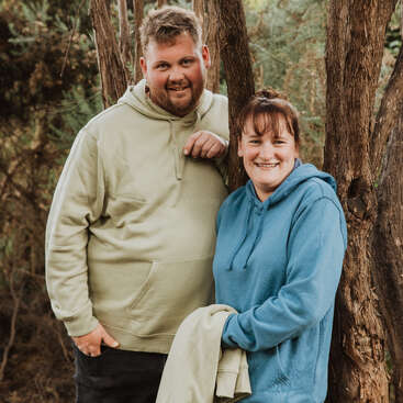 Un homme et une femme souriants se tiennent à l'extérieur près d'un arbre, tous deux vêtus de sweats à capuche. Le décor est naturel, avec de la verdure et des arbres, ce qui crée une ambiance détendue et décontractée.
