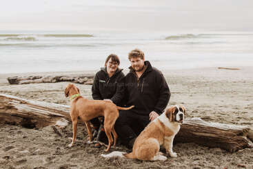 Ein Paar sitzt an einem Sandstrand neben zwei großen Baumstämmen, begleitet von zwei Hunden, während im Hintergrund unter einem bewölkten Himmel sanft die Wellen rollen.