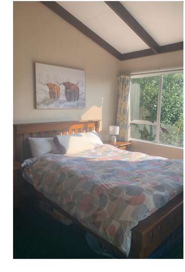 A luz do sol inunda um quarto aconchegante com uma cama de madeira, edredom estampado, mesinha de cabeceira, abajur, cortinas florais e um quadro de uma vaca das Terras Altas acima da cama. Um ambiente tranquilo e acolhedor.