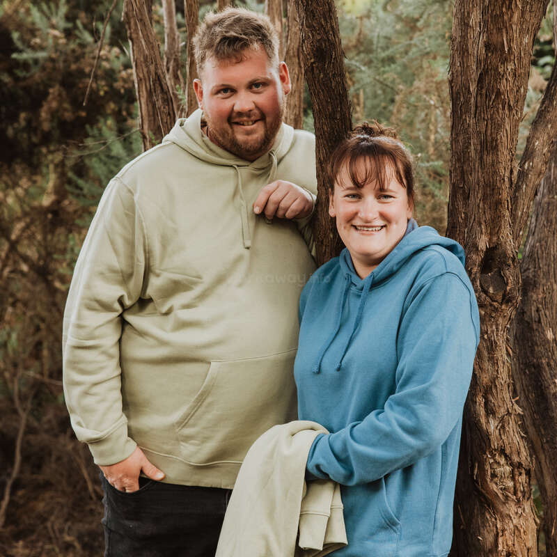 Un homme et une femme souriants se tiennent à l'extérieur près d'un arbre, tous deux vêtus de sweats à capuche. Le décor est naturel, avec de la verdure et des arbres, ce qui crée une ambiance détendue et décontractée.
