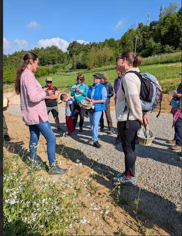 Um grupo de pessoas está ao ar livre em um dia ensolarado, ouvindo uma mulher falar. Alguns carregam cestas, cercados por vegetação, flores silvestres e céu azul.
